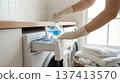 Person wearing gloves pours blue liquid into washing machine drawer in home laundry room. Neat white towels are stacked nearby. Concept of laundry services, cleaning products, household chores. 137413570