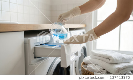 Person wearing gloves pours blue liquid into washing machine drawer in home laundry room. Neat white towels are stacked nearby. Concept of laundry services, cleaning products, household chores. Person wearing gloves pours blue liquid into washing machine drawer in home laundry room. Neat white towels are stacked nearby. Concept of laundry services, cleaning products, household chores. 137413570