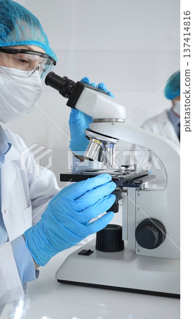 Female scientist wearing a mask and blue safety gear, using a microscope for research while a male colleague works in the background. Medicine, healthcare and science concept 137414816