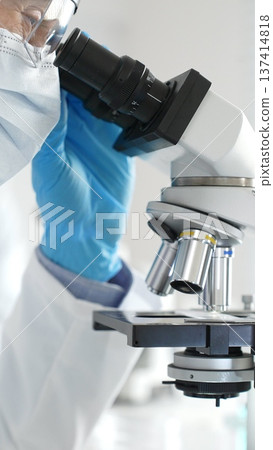 Close up of male researcher examining sample through microscope, wearing blue safety gear in modern laboratory setting. Medicine, healthcare and science concept Close up of male researcher examining sample through microscope, wearing blue safety gear in modern laboratory setting. Medicine, healthcare and science concept 137414818