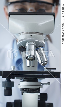 Scientist wearing lab coat and protective glasses using a modern microscope in a laboratory setting, conducting research and experiments in a scientific environment. Health care and science concept Scientist wearing lab coat and protective glasses using a modern microscope in a laboratory setting, conducting research and experiments in a scientific environment. Health care and science concept 137414837