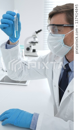 Scientist in a white lab coat, face mask, and gloves, analyzing a blue liquid in a test tube within a laboratory setting. Medicine, healthcare and science concept Scientist in a white lab coat, face mask, and gloves, analyzing a blue liquid in a test tube within a laboratory setting. Medicine, healthcare and science concept 137414845