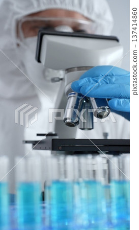 Scientific researcher or virologist wearing protective gear adjusting microscope, analyzing lab samples with precision in sterile research environment. Medicine and science concept Scientific researcher or virologist wearing protective gear adjusting microscope, analyzing lab samples with precision in sterile research environment. Medicine and science concept 137414860