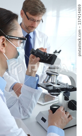 Two scientists in lab coats and safety gear conducting research, one observing through a microscope and the other holding a test tube with turquoise liquid. Medicine and science concept 137414869