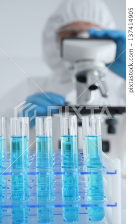 Close up of test tubes with blue liquid opposite a scientific researcher wearing protective gear tuning microscope lenses in laboratory. Medicine, healthcare and science concept Close up of test tubes with blue liquid opposite a scientific researcher wearing protective gear tuning microscope lenses in laboratory. Medicine, healthcare and science concept 137414905