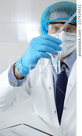 Scientist in protective gear analyzing a test tube of turquoise liquid with a pipette in a modern laboratory setting. Medicine, health care and science concept 137414931