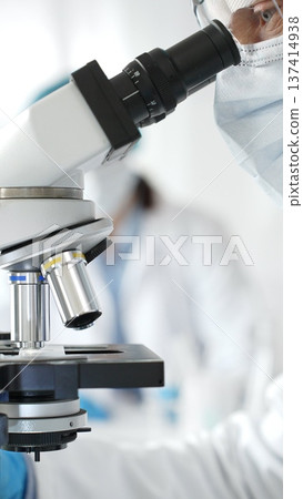 Close up of male researcher examining sample through microscope, wearing blue safety gear in modern laboratory setting. Medicine, healthcare and science concept Close up of male researcher examining sample through microscope, wearing blue safety gear in modern laboratory setting. Medicine, healthcare and science concept 137414938