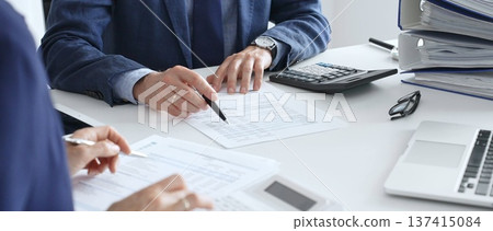 Accountants analyzing financial documents, calculating numbers together at shared workspace with laptop and office supplies surrounding them. Audit and taxes in business Accountants analyzing financial documents, calculating numbers together at shared workspace with laptop and office supplies surrounding them. Audit and taxes in business 137415084