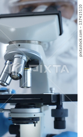 Medical virologist in a hazmat suit, mask, and gloves, diligently working at a microscope in a laboratory researching viruses. Healthcare and science concept 137415110