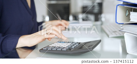 Woman calculating finances on a desk in a modern office, working on accounting, banking, and budget planning. Audit and taxes in business Woman calculating finances on a desk in a modern office, working on accounting, banking, and budget planning. Audit and taxes in business 137415112