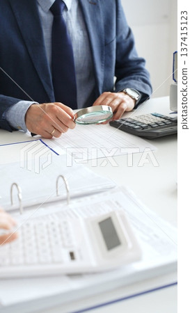 Male financial analysts examining documents, using magnifying glass and calculator while studying corporate data at workplace. Audit and taxes concept Male financial analysts examining documents, using magnifying glass and calculator while studying corporate data at workplace. Audit and taxes concept 137415123
