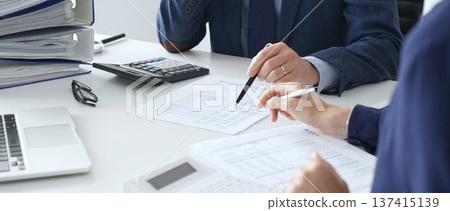 Male accountant analyzing financial documents, calculating numbers together at shared workspace with laptop and office supplies surrounding them. Audit and taxes concept Male accountant analyzing financial documents, calculating numbers together at shared workspace with laptop and office supplies surrounding them. Audit and taxes concept 137415139