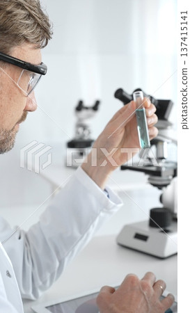 Senior male scientific researcher wearing white lab coat, analyzing blue liquid sample with tablet and microscope during medical investigation. Medicine and science concept Senior male scientific researcher wearing white lab coat, analyzing blue liquid sample with tablet and microscope during medical investigation. Medicine and science concept 137415141