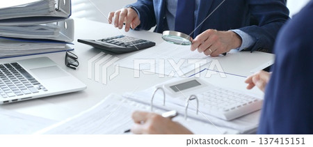 Financial analysts examining documents, using magnifying glass and calculator while studying corporate data at workplace. Audit and taxes in business Financial analysts examining documents, using magnifying glass and calculator while studying corporate data at workplace. Audit and taxes in business 137415151