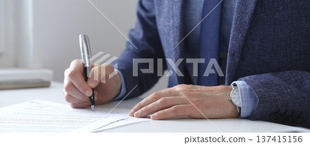 Professional businessman signing contract with silver pen, wearing tailored blue suit, business documents and digital tablet nearby, close up of hands. Business people concept 137415156