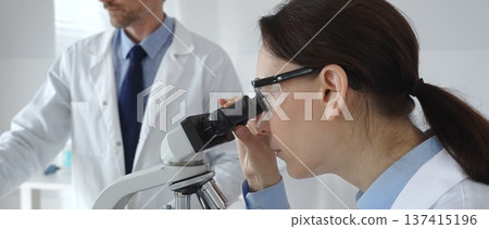Female scientist wearing protective glasses looking through a microscope during medical research in laboratory with her male colleague working in the background. Medicine and science concept Female scientist wearing protective glasses looking through a microscope during medical research in laboratory with her male colleague working in the background. Medicine and science concept 137415196
