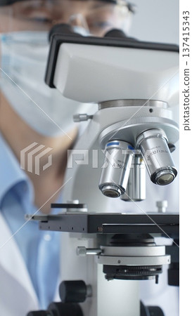 Scientist wearing lab coat and protective glasses using a modern microscope in a laboratory setting, conducting research and experiments in a scientific environment. Health care and science concept Scientist wearing lab coat and protective glasses using a modern microscope in a laboratory setting, conducting research and experiments in a scientific environment. Health care and science concept 137415343
