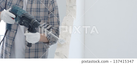 Male construction worker demolishing white wall with rotary hammer drill is wearing grey checkered shirt and protective white gloves, generating dust during renovation project, closeup view 137415347