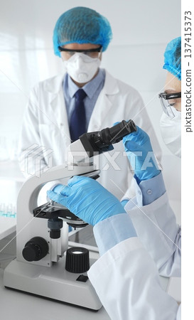 Two scientists in lab coats, face masks, and protective glasses conducting research with a microscope in a laboratory. Medicine and science concept Two scientists in lab coats, face masks, and protective glasses conducting research with a microscope in a laboratory. Medicine and science concept 137415373