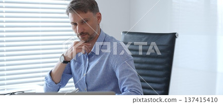 Businessman wearing blue sleek shirt is thinking and concentrating while working at the desk in a bright, modern office, demonstrating professionalism and efficiency. Business people concept 137415410
