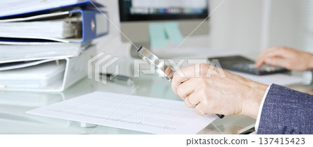 Male accountant holding magnifying glass, checking financial documents on office desk, analyzing taxes and data. Audit and taxes in business Male accountant holding magnifying glass, checking financial documents on office desk, analyzing taxes and data. Audit and taxes in business 137415423