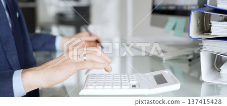 Accountant working in office, calculating finance data using black calculator and taking notes for budget planning. Audit and taxes in business Accountant working in office, calculating finance data using black calculator and taking notes for budget planning. Audit and taxes in business 137415428