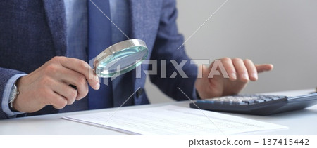 Businessman financial analyst is analyzing financial documents with a calculator and magnifying glass, ensuring accuracy and compliance in the office background. Audit and taxes in business Businessman financial analyst is analyzing financial documents with a calculator and magnifying glass, ensuring accuracy and compliance in the office background. Audit and taxes in business 137415442