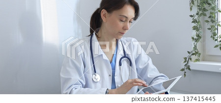 Female doctor wearing lab coat and stethoscope using digital tablet while sitting at desk in medical office, browsing patient information or conducting online consultation. Medicine concept 137415445