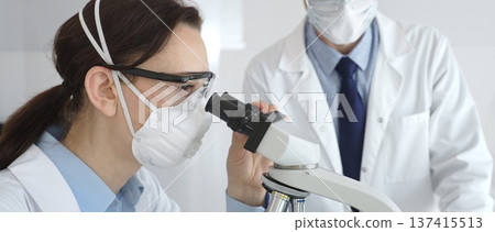 Two scientists in lab coats and safety gear conducting research, one observing through a microscope and the other holding a test tube with turquoise liquid. Medicine and science concept 137415513