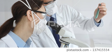 Two scientists in lab coats and protective gear conduct research in a sterile laboratory: one observes through a microscope, and the other holds a test tube with turquoise liquid. Medicine and science 137415554