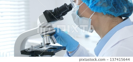 Female scientist analyzing research sample through microscope, wearing protective lab gear for medical investigation. Laboratory and science concept 137415644