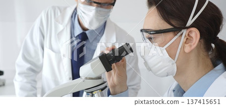 Two scientists in lab coats and safety gear conducting research, one observing through a microscope and the other holding a test tube with turquoise liquid. Medicine and science concept 137415651