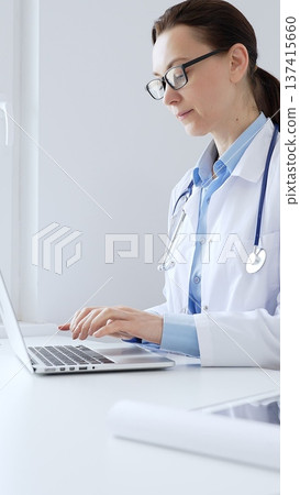 Female doctor is typing on a laptop in a bright medical office, showcasing healthcare technology and medical documentation. Medicine and health care concept 137415660