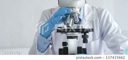 Scientist wearing protective gear carefully adjusting microscope lens, analyzing medical research samples with precision in sterile laboratory environment Scientist wearing protective gear carefully adjusting microscope lens, analyzing medical research samples with precision in sterile laboratory environment 137415662