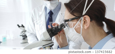 Two scientists in lab coats and safety gear conducting research, one observing through a microscope and the other holding a test tube with turquoise liquid. Medicine and science concept Two scientists in lab coats and safety gear conducting research, one observing through a microscope and the other holding a test tube with turquoise liquid. Medicine and science concept 137415682