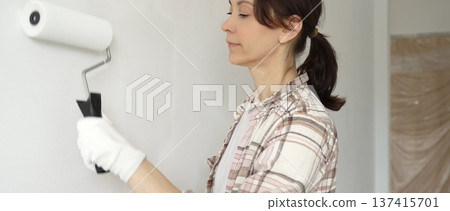 Middle aged woman wearing gloves is skillfully applying paint to a white wall with a paint roller in an apartment undergoing renovation, transforming the space with her creative touch Middle aged woman wearing gloves is skillfully applying paint to a white wall with a paint roller in an apartment undergoing renovation, transforming the space with her creative touch 137415701