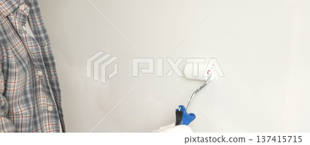 Male painter's gloved hand gripping paint roller, spreading white paint across wall during home interior transformation with professional precision, close-up view. Renovation concept Male painter's gloved hand gripping paint roller, spreading white paint across wall during home interior transformation with professional precision, close-up view. Renovation concept 137415715