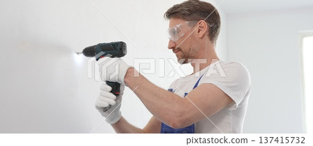 Man professional construction worker in a blue jumpsuit, safety goggles, and work gloves is screwing a screw into the wall using a cordless screwdriver, home renovation 137415732