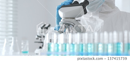 Male doctor in protective suit and gloves using microscope while analyzing samples while conducting research in sterile laboratory. Close-up of test tubes with blue liquid Male doctor in protective suit and gloves using microscope while analyzing samples while conducting research in sterile laboratory. Close-up of test tubes with blue liquid 137415739