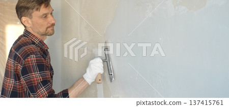 Male construction worker wearing gloves is carefully applying plaster to a wall using a hand tool, contributing to a home renovation or construction project Male construction worker wearing gloves is carefully applying plaster to a wall using a hand tool, contributing to a home renovation or construction project 137415761