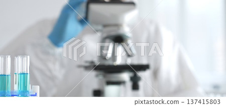 Male medical scientist in protective suit using microscope while analyzing samples, conducting research in laboratory. Close-up of test tubes with blue liquid Male medical scientist in protective suit using microscope while analyzing samples, conducting research in laboratory. Close-up of test tubes with blue liquid 137415803