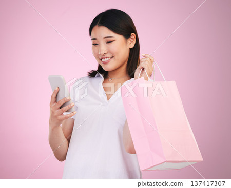Happy asian woman, phone and shopping bag in payment, purchase or buying against a pink studio background. Female person or shopper on mobile smartphone banking app in ecommerce, sale or discount Happy asian woman, phone and shopping bag in payment, purchase or buying against a pink studio background. Female person or shopper on mobile smartphone banking app in ecommerce, sale or discount 137417307