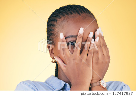 Shy, black woman and hands on face peeping in studio isolated on yellow background. Portrait, fingers and African person embarrassed, scared at danger or fear at horror, social anxiety and spy hiding 137417987