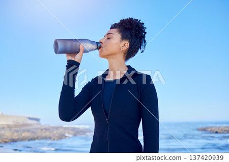 Fitness, woman and drinking water at beach for running, health or morning cardio on blue sky by an ocean. Exercise, hydration and thirsty lady runner with sports liquid after training or sea workout Fitness, woman and drinking water at beach for running, health or morning cardio on blue sky by an ocean. Exercise, hydration and thirsty lady runner with sports liquid after training or sea workout 137420939