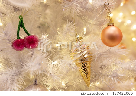 Cute pastel Christmas ornaments, including a pink cherry, golden ice cream cone, and peach bauble, hanging on a white tree with warm glowing lights. A whimsical and festive holiday close-up. 137421060