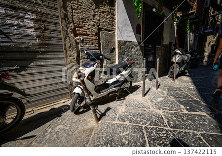 Narrow cobblestone street in historic Naples, Italy, featuring parked scooters and weathered stone buildings. A white scooter is locked to a metal post. Shuttered doors and rough-textured walls Narrow cobblestone street in historic Naples, Italy, featuring parked scooters and weathered stone buildings. A white scooter is locked to a metal post. Shuttered doors and rough-textured walls 137422115