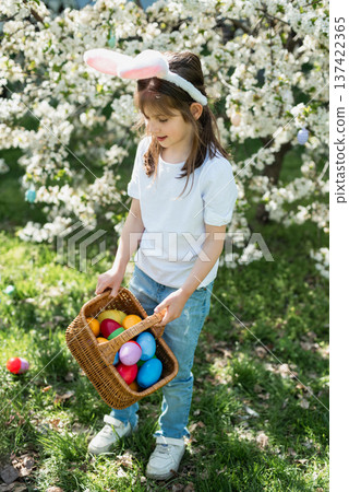 Easter egg hunt. Little girl with bunny ears holding decorative Easter egg and wicker basket under blooming spring tree. Outdoor Easter celebration. joyful childhood moment outdoors. 137422365