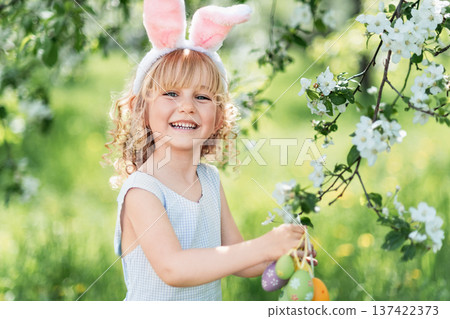 Outdoor Easter celebration, childhood joy and seasonal tradition. Little girl with bunny ears holding decorative Easter egg and wicker basket under blooming spring tree. 137422373