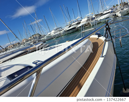 The bow of a sailing yacht with shiny railings. Many yachts are docked in the harbor under a clear blue sky. Masts rise in the background, indicating a busy marina. The bow of a sailing yacht with shiny railings. Many yachts are docked in the harbor under a clear blue sky. Masts rise in the background, indicating a busy marina. 137422667