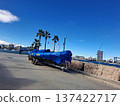 A large motorboat is parked on a trailer at a Mediterranean pier. The boat is covered with a blue tarp. Palm trees line the area, and the sky is clear above the water. 137422717